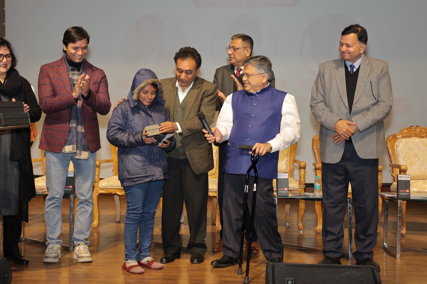 A visually impaired woman receives her SMARTON Kit on stage at a distribution event, as dignitaries applaud